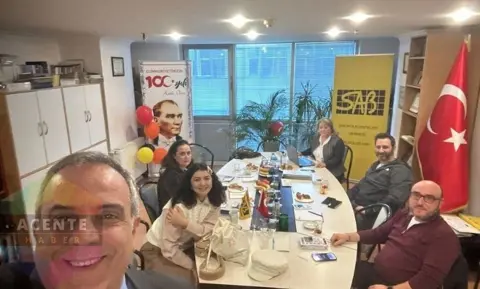 SAB ofisinde altı kişi masa başında selfie çekiliyor, arkada Atatürk ve balonlar